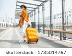 Young female traveler walking with a yellow suitcase at the modern transport stop outdoors, back view. Concept of an urban transportation and travel