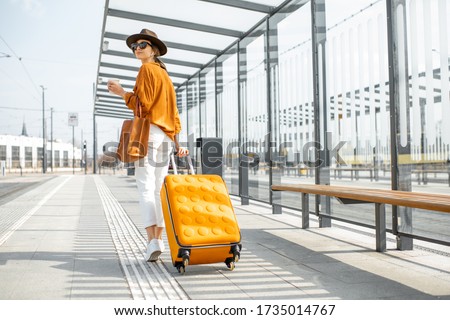 Young female traveler walking with a yellow suitcase at the modern transport stop outdoors, back view. Concept of an urban transportation and travel