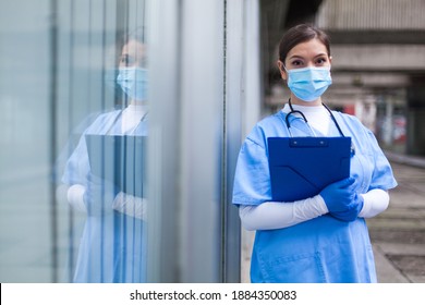 Young Female UK EMS Doctor In Front Of Healthcare ICU Facility,wearing Protective Face Mask Holding Medical Patient Health Check Form,reflection In The Glass Window,Coronavirus Pandemic Crisis 