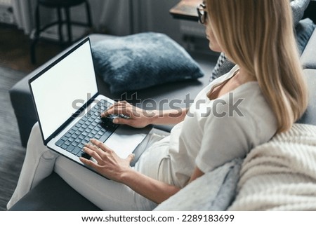 young female using laptop computer for browsing online website, sitting on comfortable couch and typing on keyboard, over shoulder camera shot 