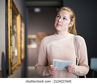 Young Female Visitor With Guide Book Looking At Exhibition In Museum