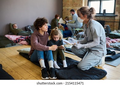 Young female volunteer in medical uniform consulting refugee with child while sitting on squats on their sleeping place in front
