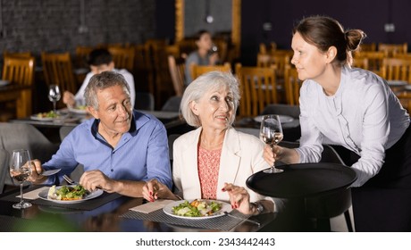 Young female waiter serves mature couple of positive spouses in restaurant. Hostess serves puts on table order for dinner dishes of national Mediterranean cuisine - Powered by Shutterstock