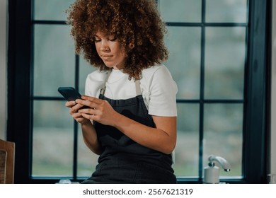 Young female waitress wearing apron, using smartphone in restaurant kitchen next to window, taking a break, texting, reading messages, or using app