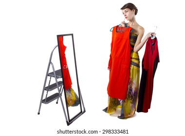 young female wearing clothes in a fitting room with a mirror. she is choosing between multiple dresses and indecisive.  She is holding a red dress.