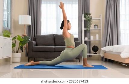 Young female wearing green sportswear and practicing morning yoga routine at home, standing in Low Crescent Lunge pose or Anjaneyasana to stretches thighs and groins on blue mat in bedroom.  - Powered by Shutterstock