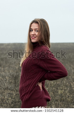 Young female wearing stylish sweater and 
leggings with a pattern half-turned and look back with a smile outdoors in the wild plain field at autumn foggy cold weather