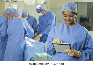 Young female woman nurse doctor surgeon using tablet computer with her interracial surgical team working on a patient in hospital surgery operating theater in the background