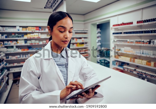 Young female woman working\
in pharmacy standing behind counter wearing labcoat using digital\
tablet