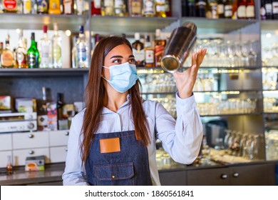 Young Female Worker At Bartender Desk In Restaurant Bar Preparing Coctail With Shaker. Beautiful Young Woman Behind Bar Wearing Protective Face Mask