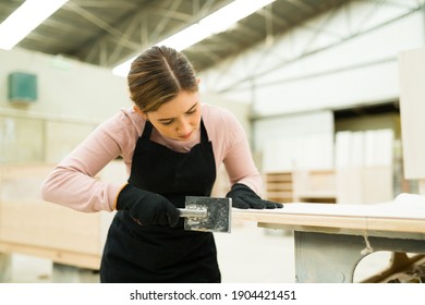 Young Female Worker And Carpenter Working With A Chisel Tool Around The Edges Of A Wooden Table Inside A Woodshop