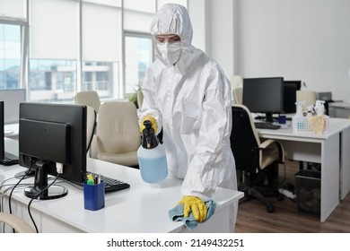 Young Female Worker Of Cleaning Service In Hazmat Suit Wiping Surfaces Of Desks And Computer Equipment In Modern Openspace Office