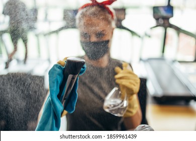 Young Female Worker Disinfecting Cleaning And Weeping Expensive Fitness Gym Equipment With Alcohol Sprayer And Cloth. Coronavirus Global World Pandemic And Health Protection Safety Measures.