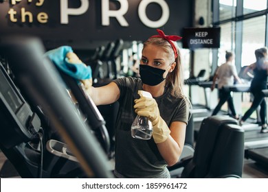 Young Female Worker Disinfecting Cleaning And Weeping Expensive Fitness Gym Equipment With Alcohol Sprayer And Cloth. Coronavirus Global World Pandemic And Health Protection Safety Measures.