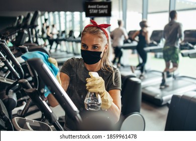 Young Female Worker Disinfecting Cleaning And Weeping Expensive Fitness Gym Equipment With Alcohol Sprayer And Cloth. Coronavirus Global World Pandemic And Health Protection Safety Measures.