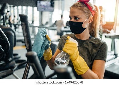 Young female worker disinfecting cleaning and weeping expensive fitness gym equipment with alcohol sprayer and cloth. Coronavirus global world pandemic and health protection safety measures. - Shutterstock ID 1949425078