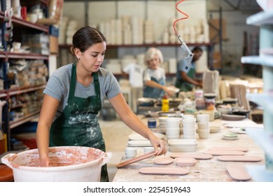 Young female worker in pottery factory lowers fired clay plates and cups into a container with glaze