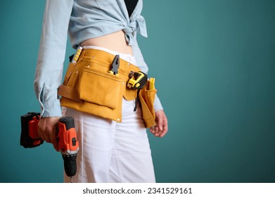 Young female worker with tool belt holding an electric screwdriver in hands isolated on background. Pretty caucasian female with tools planning new project. Portrait in studio. - Powered by Shutterstock