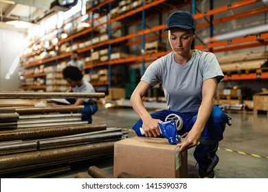 Young Female Worker Using Tape Dispenser Gun While Packing Cardboard Boxes For The Shipment In Distribution Warehouse. 