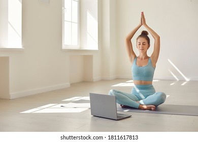 Young female yoga coach in front of computer hosting webinar. Young woman with laptop sitting cross legged learning to meditate following instructions from online video lesson - Powered by Shutterstock