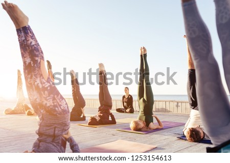 Young female yoga instructor teaching and correct a handstand pose, salamba sirsasana exercise for a group of sporty people practicing outdoor, working out, teacher helping student to master