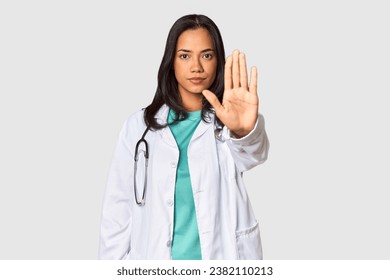 Young Filipina doctor with stethoscope in studio standing with outstretched hand showing stop sign, preventing you. - Powered by Shutterstock