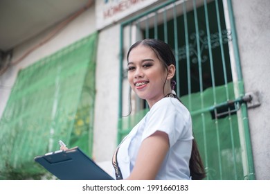 A Young Filipina Nurse Or Nursing Aide In Front Of A Small Rural Clinic In A Remote Provincial Area. NGO Or Barangay Medical Staff.