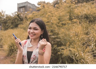 A Young Filipina Woman Blissful And Happy While Holding Her Phone. Reacting After Passing Her Board Exams Or Accepted To A New Job.