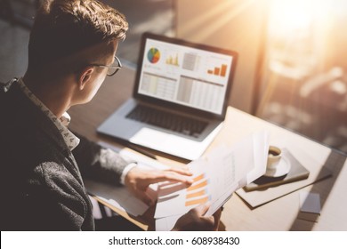 Young finance market analyst in eyeglasses working at sunny office laptop while sitting at wooden table Businessman analyze document in his hands Graphs   diagramm notebook screen Blurred