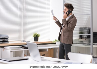 Young Financial Advisor In Brown Jacket Standing At Window Closed With Blinds And Consulting On Taxes By Phone