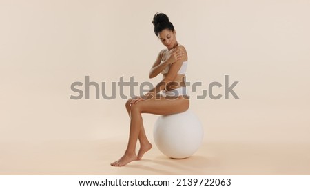 Young fit African American woman with bun hairstyle in white underwear strokes shoulder and arm, sitting on big white ball on beige background | Body care commercial concept