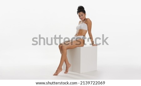 Young fit African American woman with dark curly hair in bun in white underwear sitting on white cube podium on white background | Skin care concept