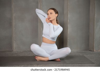 Young fit athletic woman in white sportswear sitting on yoga mat in lotus pose, holding hands near face and looking ahead.

Sporty woman working out, warming up or doing exercise on a grey background.
