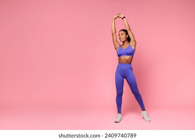 Young Fit Black Lady In Purple Fitwear Stretching Raised Arms Standing Over Pink Background, Studio Shot. African American Sportswoman Exercising And Warming Up Before Workout. Empty Space - Powered by Shutterstock