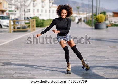 Young fit black woman on roller skates riding outdoors on urban street. Smiling girl with afro hairstyle rollerblading on sunny day