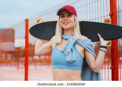 Young Fit Blonde Smiling Woman In Sportswear And Red Cap Holding Skate Behind Her Head With Hands Stands Outside, Looking Away With Confident Smile. Skateboarding Concept