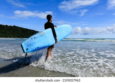 Young Fit And Firm Body Teenage Guy Walking Into Sea Holding Soft Board Ready For Extreme Sports Surfing On Beach Holidays On Clear Day, Blue Skies. Concept For Teen Hobby And Adventure In Vacation.