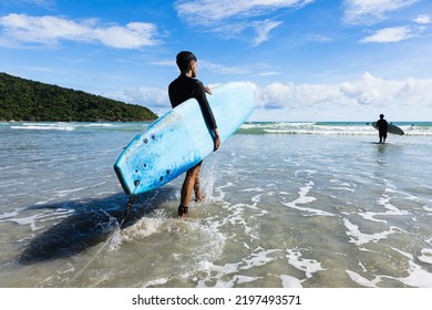 Young Fit And Firm Body Teenage Guy Walking Into Sea Holding Soft Board Ready For Extreme Sports Surfing On Beach Holidays On Clear Day, Blue Skies. Concept For Teen Hobby And Adventure In Vacation.