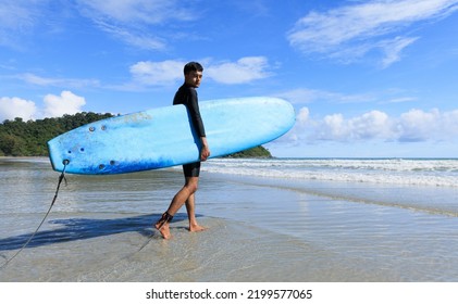 Young Fit And Firm Body Teenage Guy Walking Into Sea Holding Soft Board Ready For Extreme Sports Surfing On Beach Holidays And Looking To Camera. Concept For Teen Hobby And Adventure In Vacation.