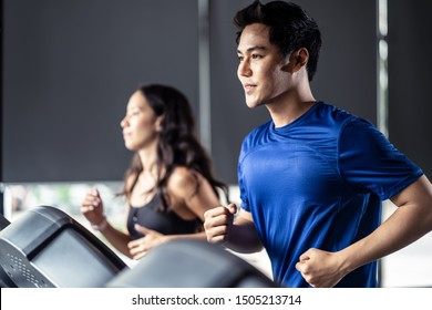 Young Fit Handsome Asian Man And Beautiful Asian Woman Running On Treadmill Or Running Machine In Modern Fitness Gym. Seen From Side View While They Focusing On Running. Workout In Gym And Fitness.