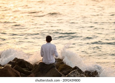 Young Fit Man In A White Linen Shirt Sitting Alone On The Rocks By The Ocean At Sunset Hour. Mental Health, Relaxation, Burnout Concept.