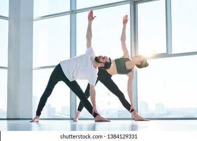 Young fit man and woman in activewear raising right arms and looking at them while side-bending in gym during yoga workout - Powered by Shutterstock