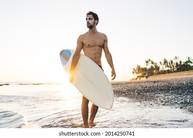 Young Fit Shirtless Male Surfer With Sunblock On Face And Surfboard Under Arm Walking Along Pebble Beach In Tide Waters On Sunny Day