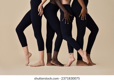 Young fit sporty diverse women wearing sportswear cloth black leggings fitness outfit pants standing in row on beige, legs feet close up shot. Multicultural girls four models isolated on background.
