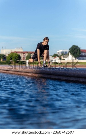 Young, fit and sporty girl in black clothes stretching after the workout near the public fountain in the urban city park. Fitness, sport, urban jogging and healthy lifestyle concept.