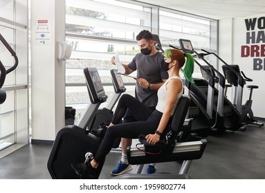 Young fit sporty woman with her personal fitness trainer exercising on seated bike. Keeping social distance and wearing protective face masks. Notices sowing pandemic rules - Powered by Shutterstock