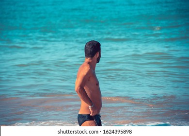 Young, Fit, Tanned And Handsome Man In Black Bathing Suit With Hands On Hist Hips Looking At An Amazing Blue Tropical Sea. Vacation Concept.