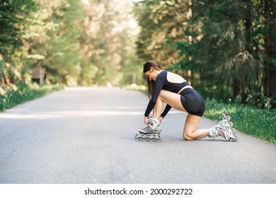 Young Fit Woman Kneeling And Fastens Roller Skates In Woods On Asphalt Road In Nature. Wearing Black Open Crop Top And Sporty Shorts.