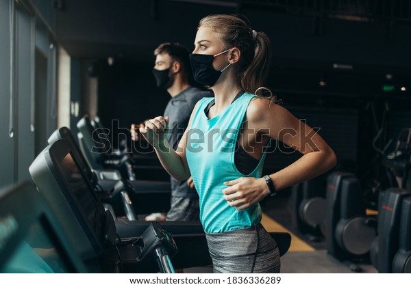 Young fit woman and man\
running on treadmill in modern fitness gym. They keeping distance\
and wearing protective face masks. Coronavirus world pandemic and\
sport theme.