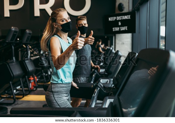Young fit woman and man\
running on treadmill in modern fitness gym. They keeping distance\
and wearing protective face masks. Coronavirus world pandemic and\
sport theme.
